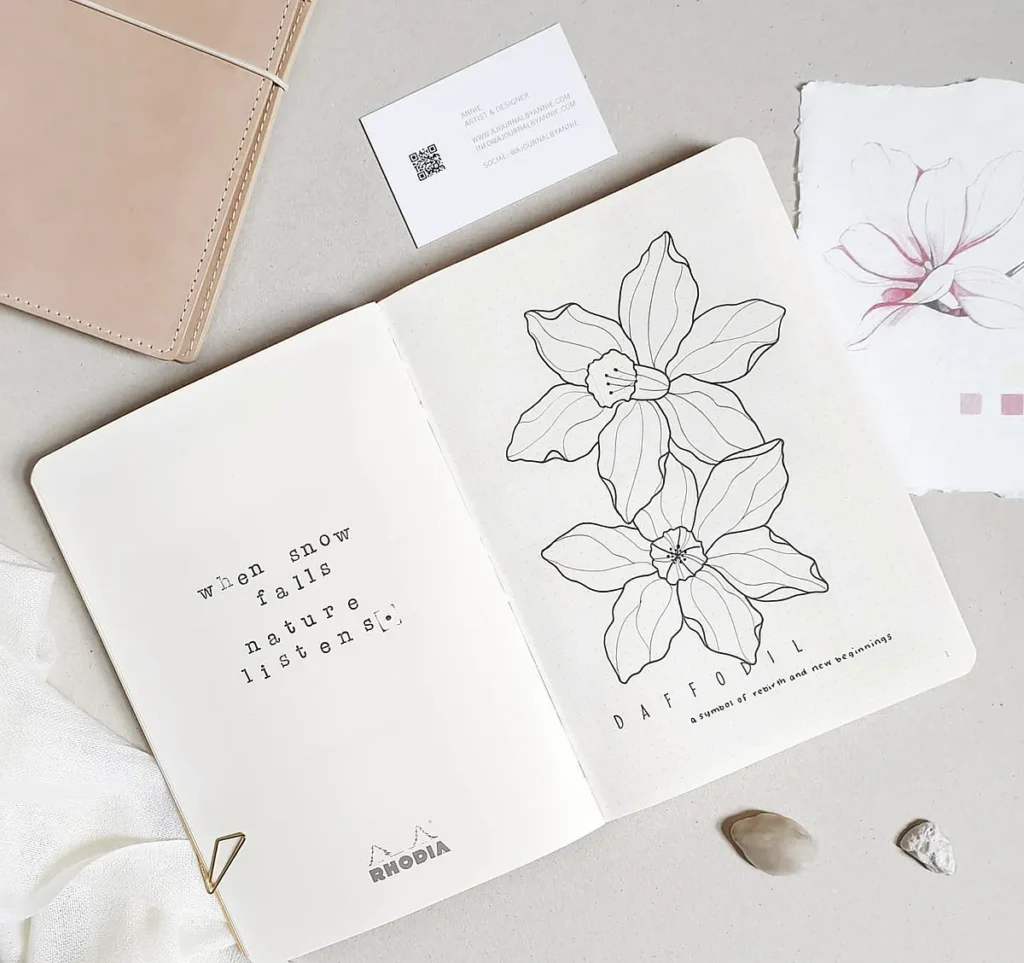 Open notebook with daffodil sketch and quote when snow falls, nature listens. Aesthetic workspace with cards and stones.