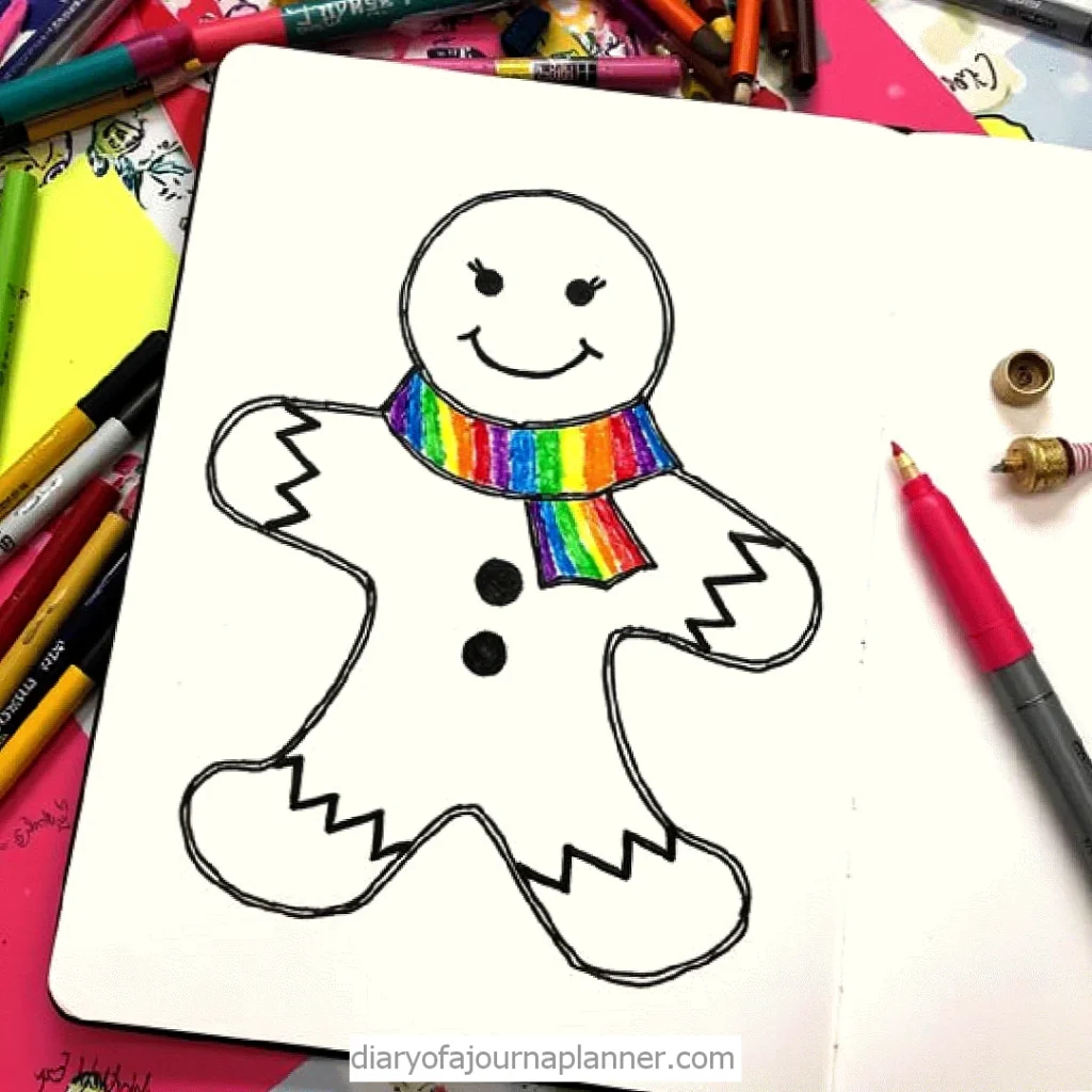 Gingerbread man drawing with a rainbow scarf in a sketchbook, surrounded by colorful pencils and pens.