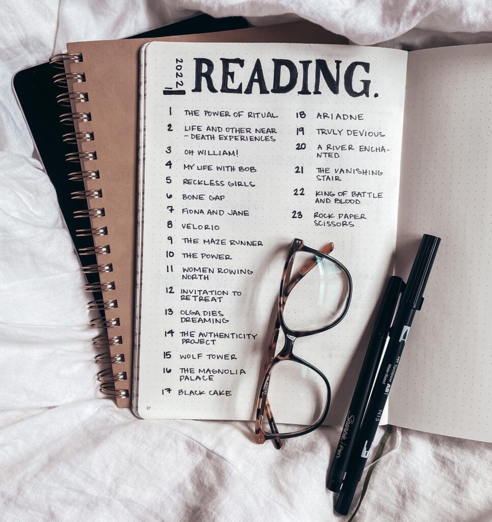 A journal with a 2022 reading list, glasses, and pens on a white fabric background.