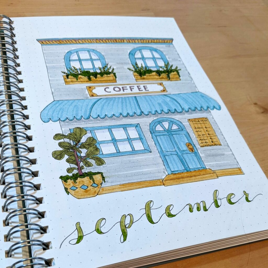 Illustrated café with blue awning and plant, titled September on spiral notebook page.