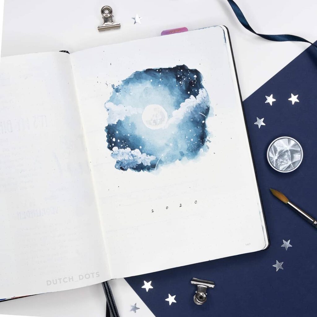 Open journal with watercolor moon and starry sky painting, surrounded by art supplies and star accents.