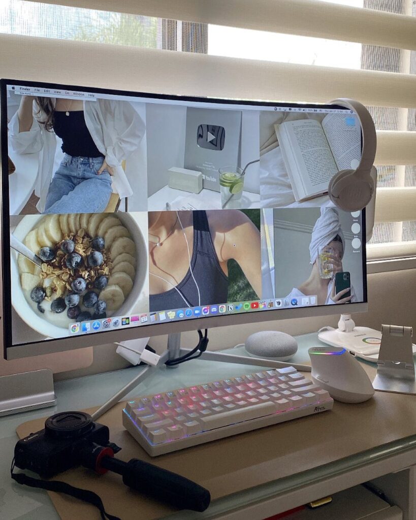 Computer monitor showing lifestyle images, white keyboard, and camera on desk, with headphones hanging on the side.