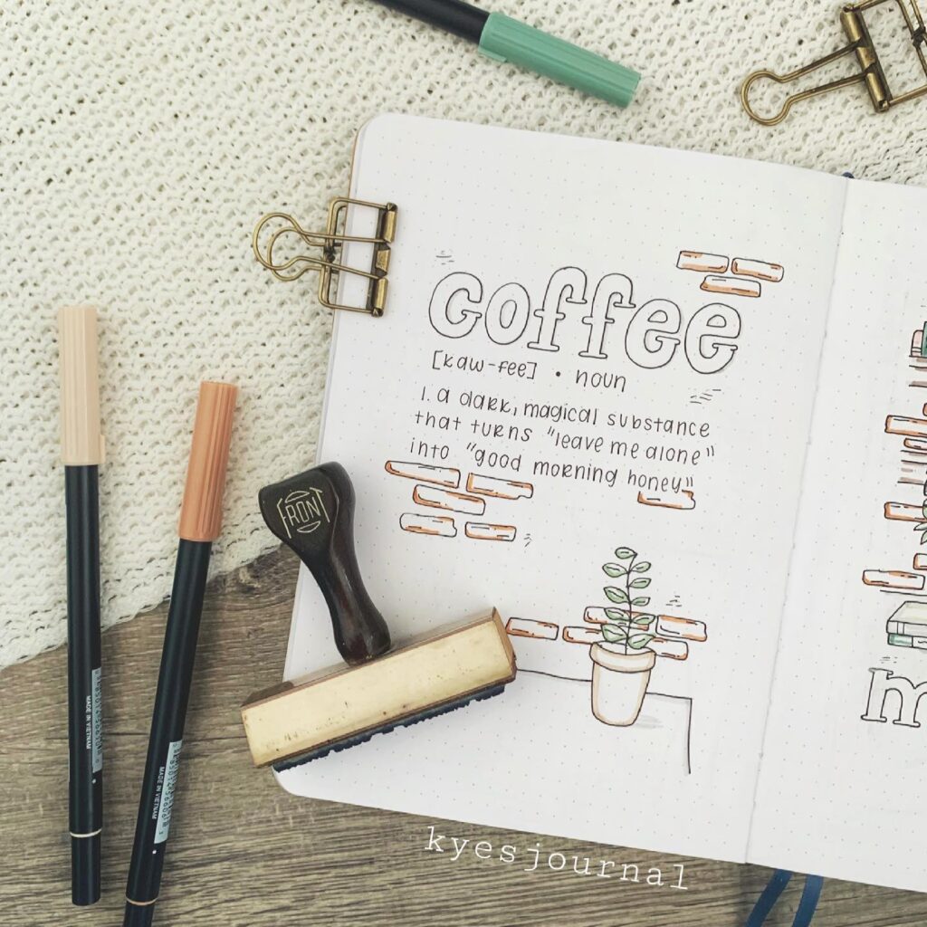 Bullet journal page with a coffee definition, surrounded by pens, stamp, and clips on a textured background.