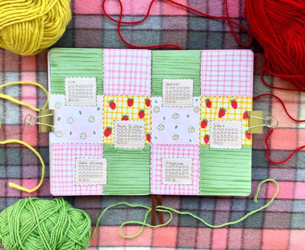 Colorful bullet journal page with strawberry and floral designs, surrounded by yarn on a plaid blanket.