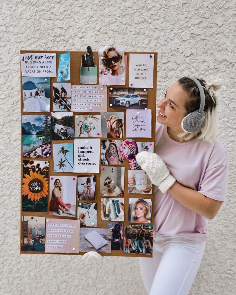 Person holding a motivational vision board with quotes and images, wearing earmuffs and mittens against a textured wall.