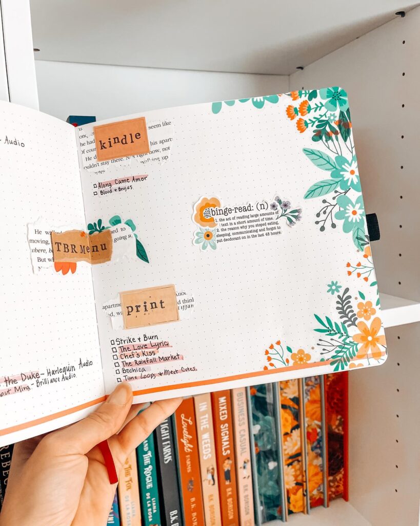 Hand holding a colorful journal with reading lists, floral designs, and bookshelf in the background.