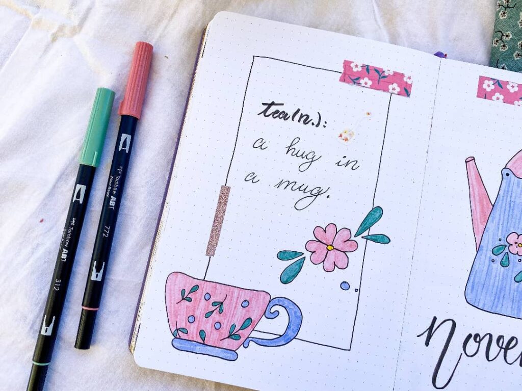 Notebook page with a teacup doodle and text a hug in a mug, alongside two colorful markers.