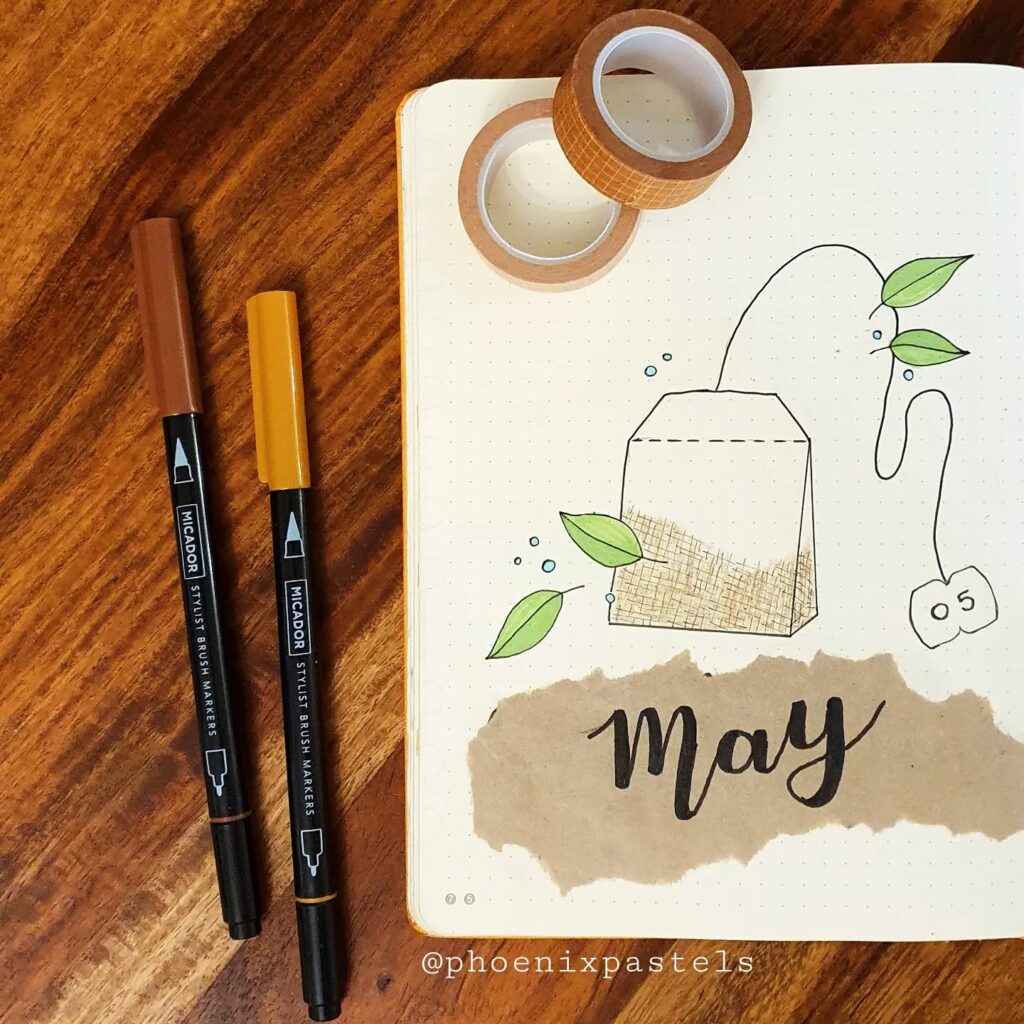 Bullet journal layout for May with tea bag doodle, brush pens, and washi tape on wooden table.