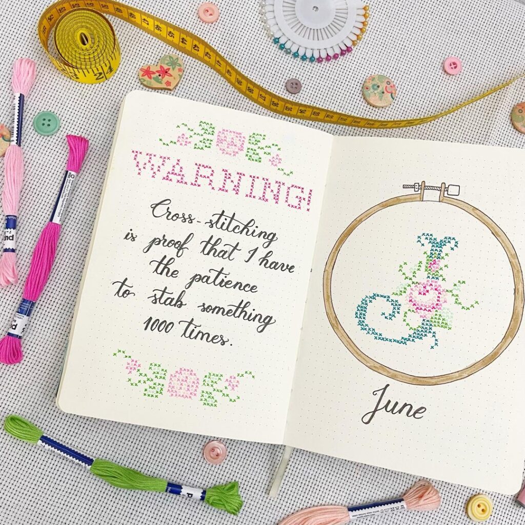 Open notebook with cross-stitching quotes and floral embroidery hoop, surrounded by colorful thread and sewing tools.