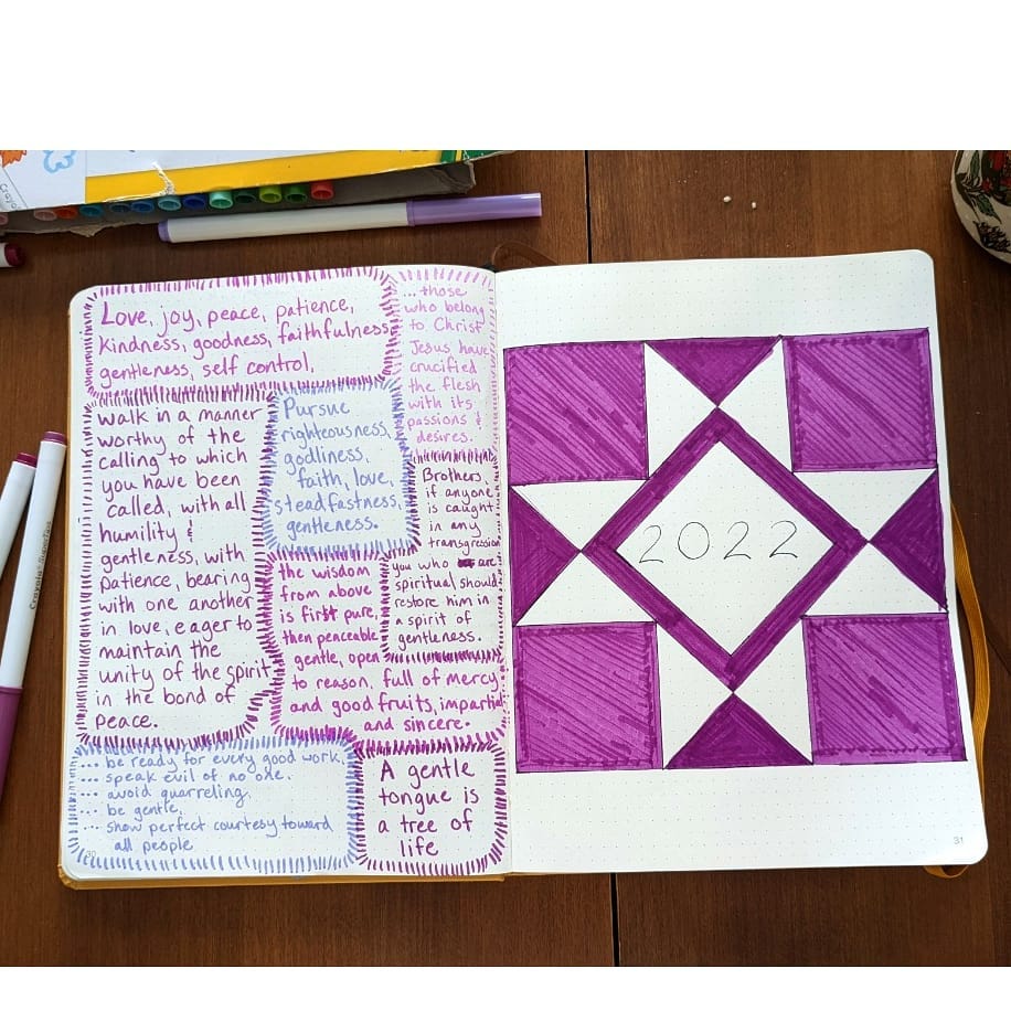 Open journal with 2022 and motivational words in purple, surrounded by pens and a cup on a wooden table.