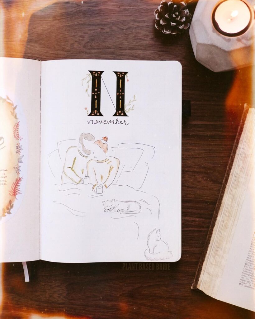 Cozy November bullet journal spread with hand-drawn couple and cats, alongside a lit candle and an open book.