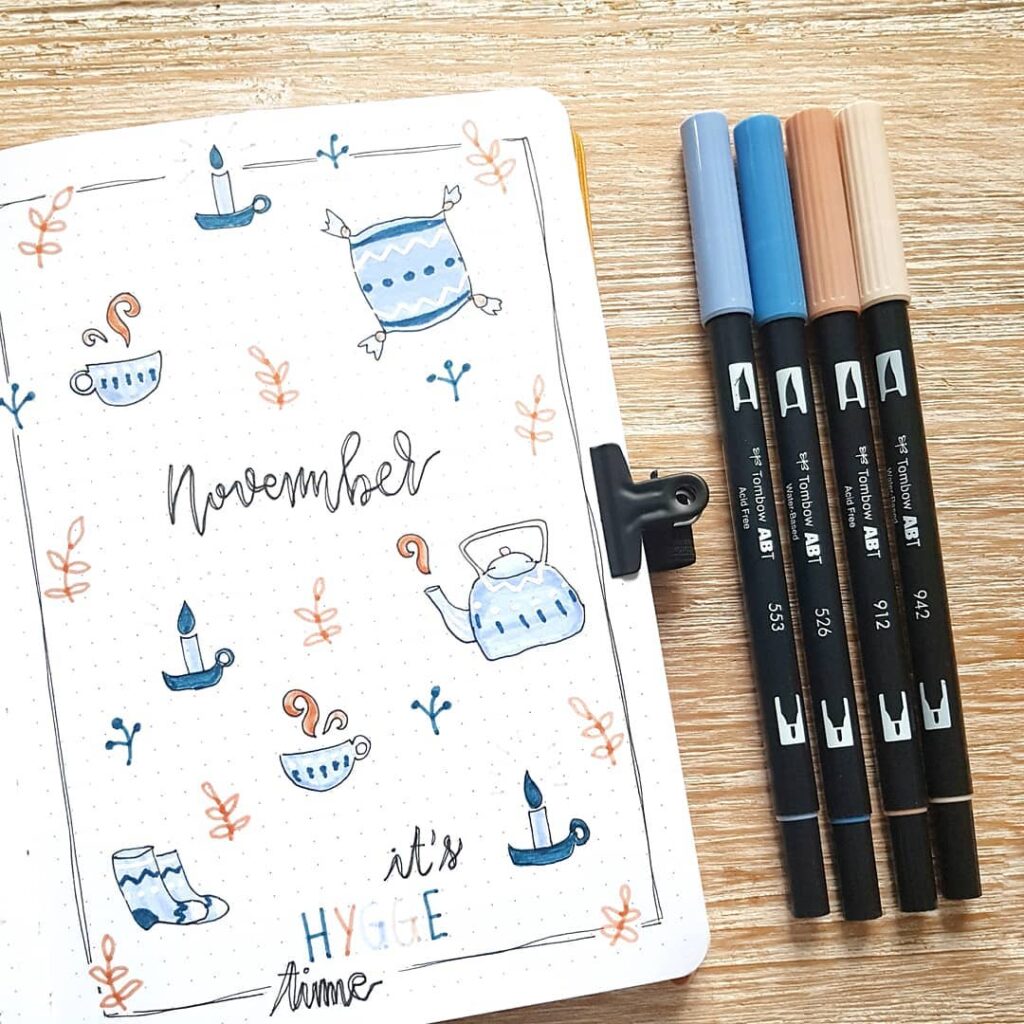 Bullet journal spread for November with cozy hygge theme, featuring candles, kettle, cup, sock, and brush pens.