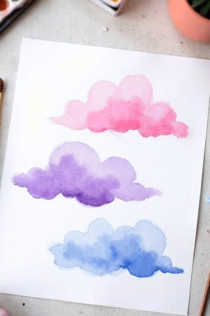 Watercolor painting of pink, purple, and blue clouds on paper, surrounded by art supplies.