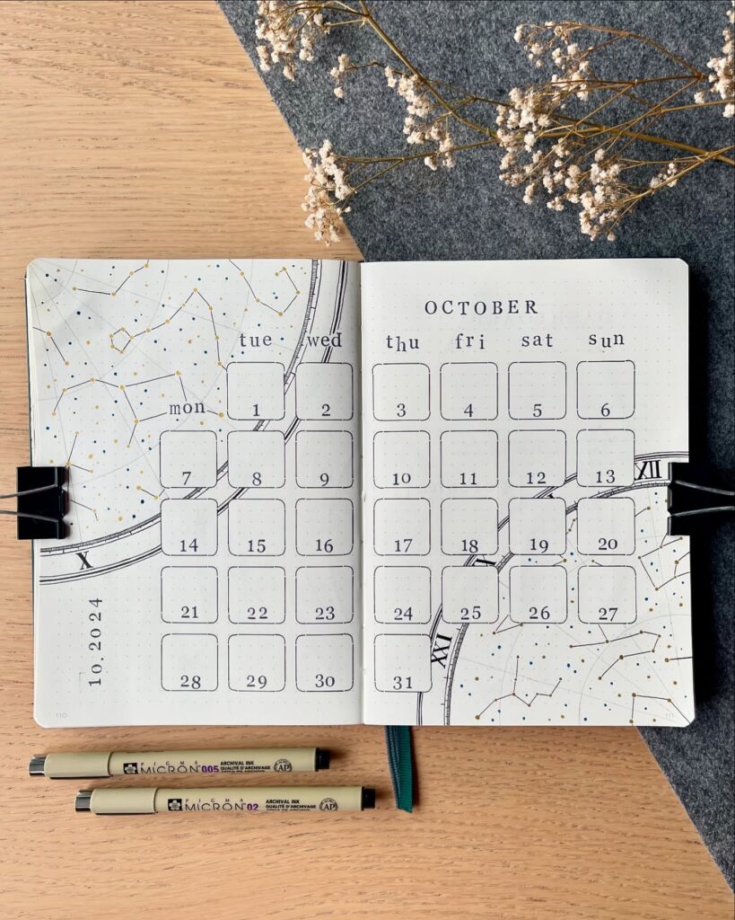October bullet journal spread with calendar layout, pens, and dried flowers on a wooden desk.