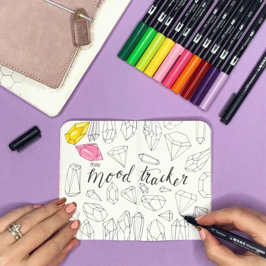 Bullet journal mood tracker with colorful pens on purple background, featuring geometric gem designs.