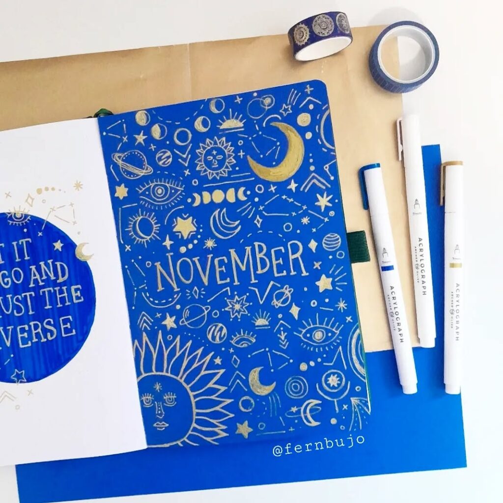 Blue and gold November bullet journal page with celestial designs, washi tape, and pens.