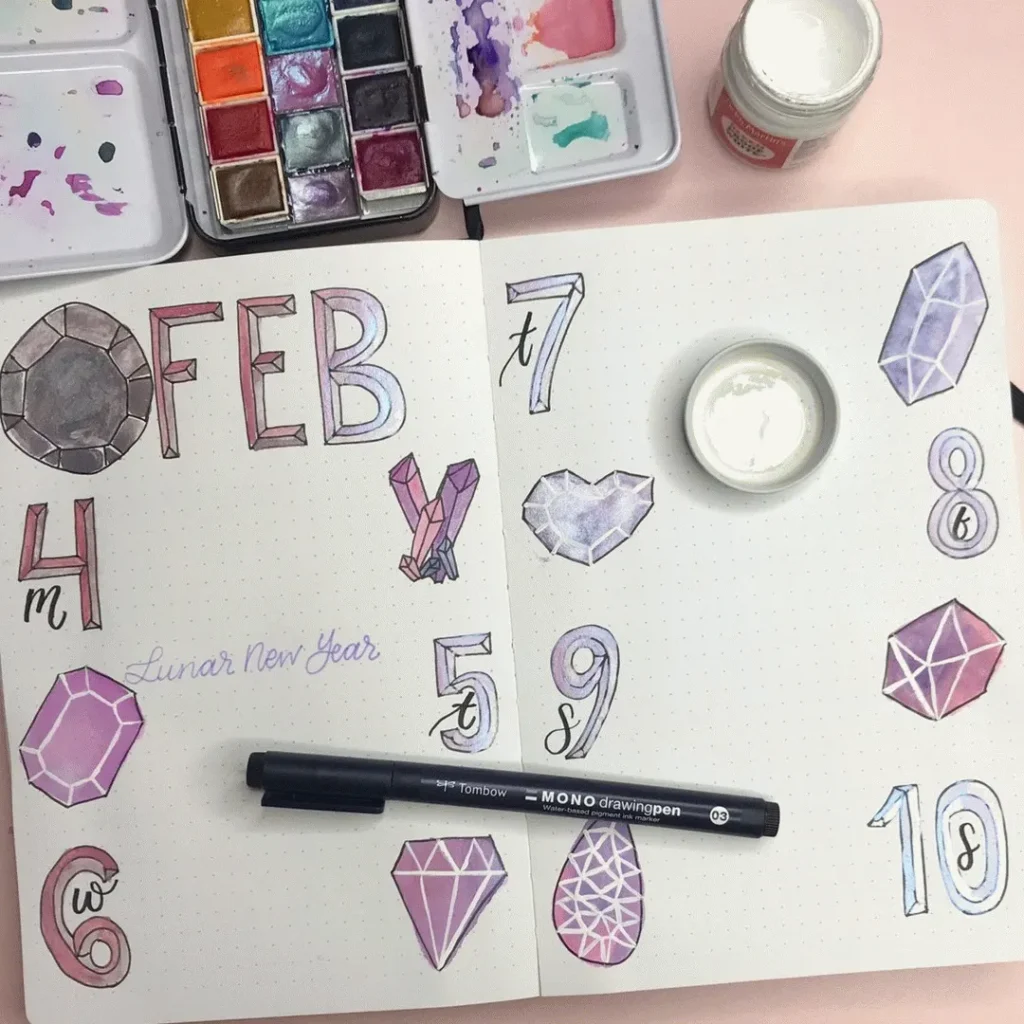 Bullet journal with February theme, decorated with watercolor gemstones and designs, Tombow pen, and paint palette.