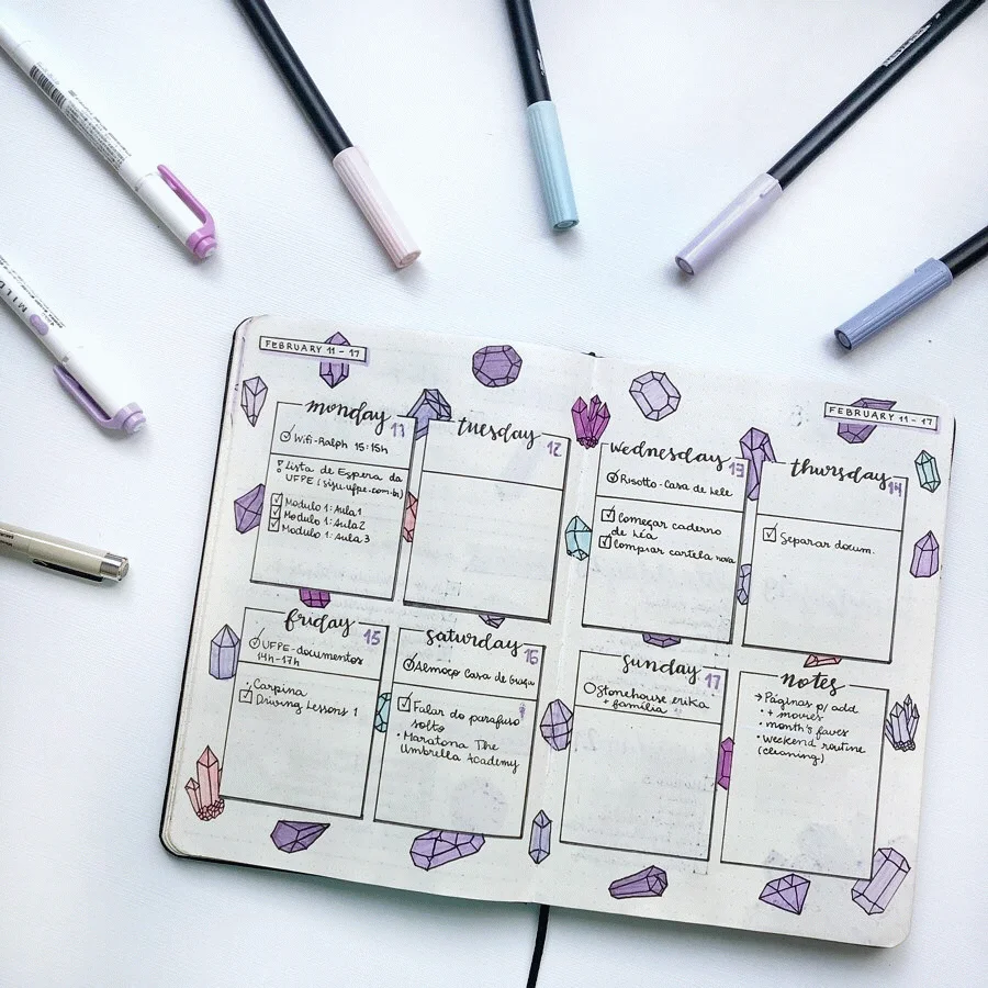 Decorative bullet journal layout with colorful gemstones and weekly planner notes surrounded by pastel pens on a white surface.