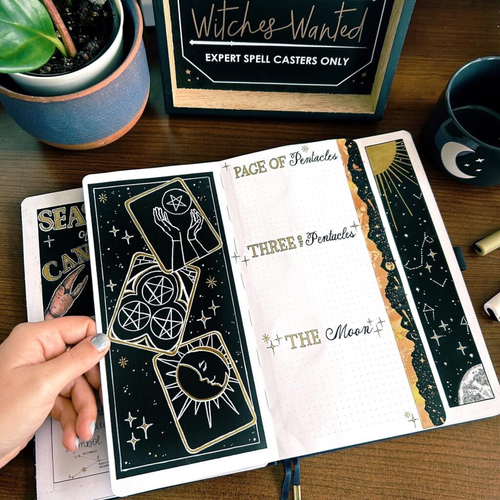 Hand holding tarot card-themed journal with mystical designs, next to plants and a mug on a wooden table.