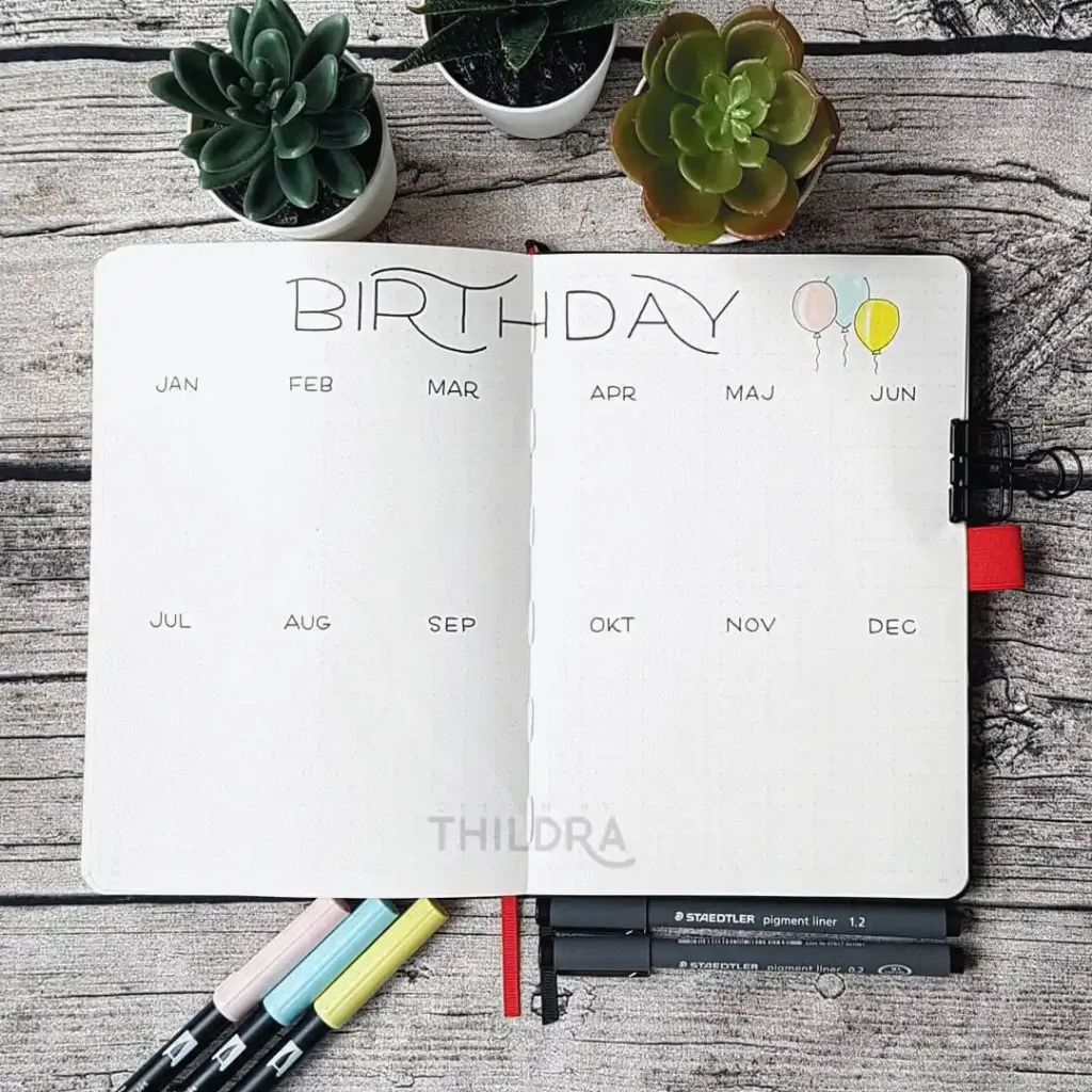 Bullet journal open to a birthday calendar page with colorful markers and succulents on a wooden background.