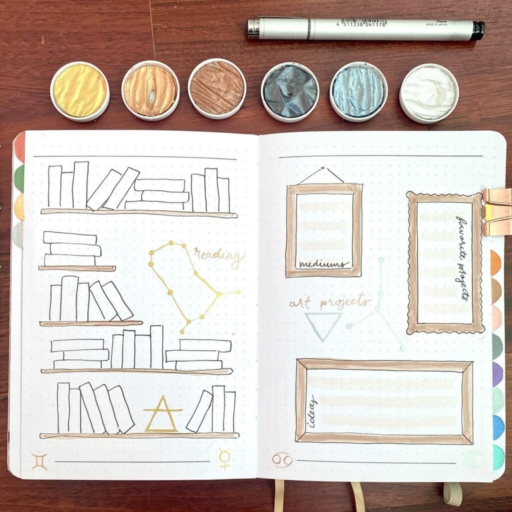 Bullet journal with artistic book sketches, frames, and metallic paint cups on a wooden desk.