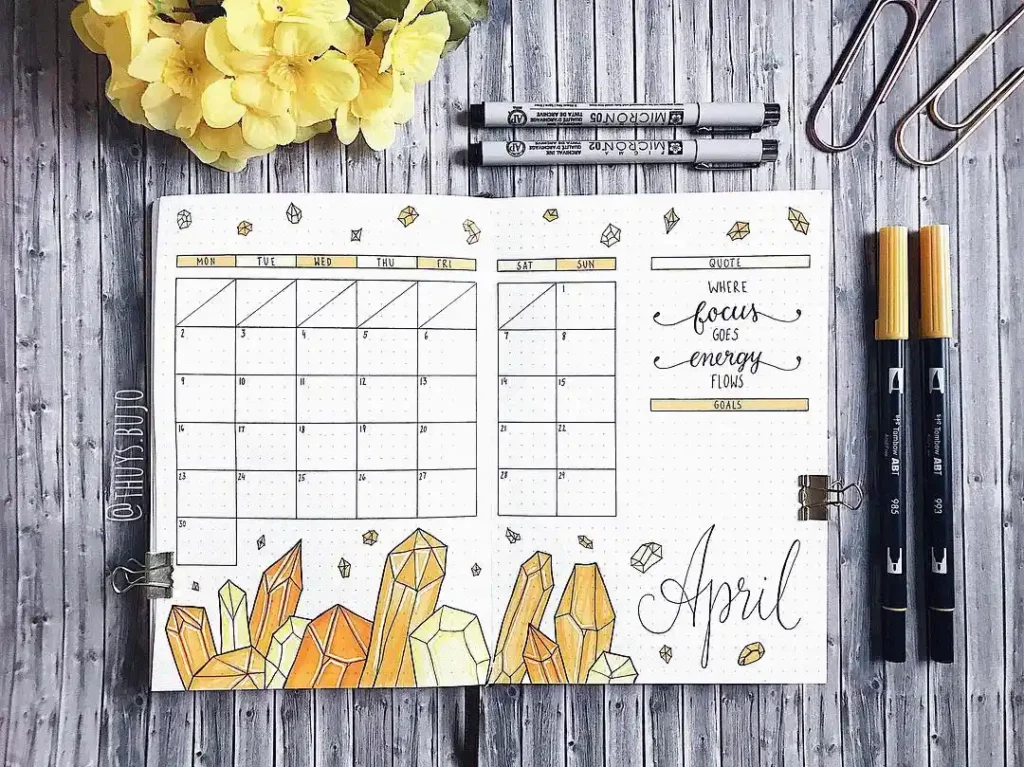 April bullet journal spread with crystals, yellow flowers, markers, and motivational quote on wooden background.