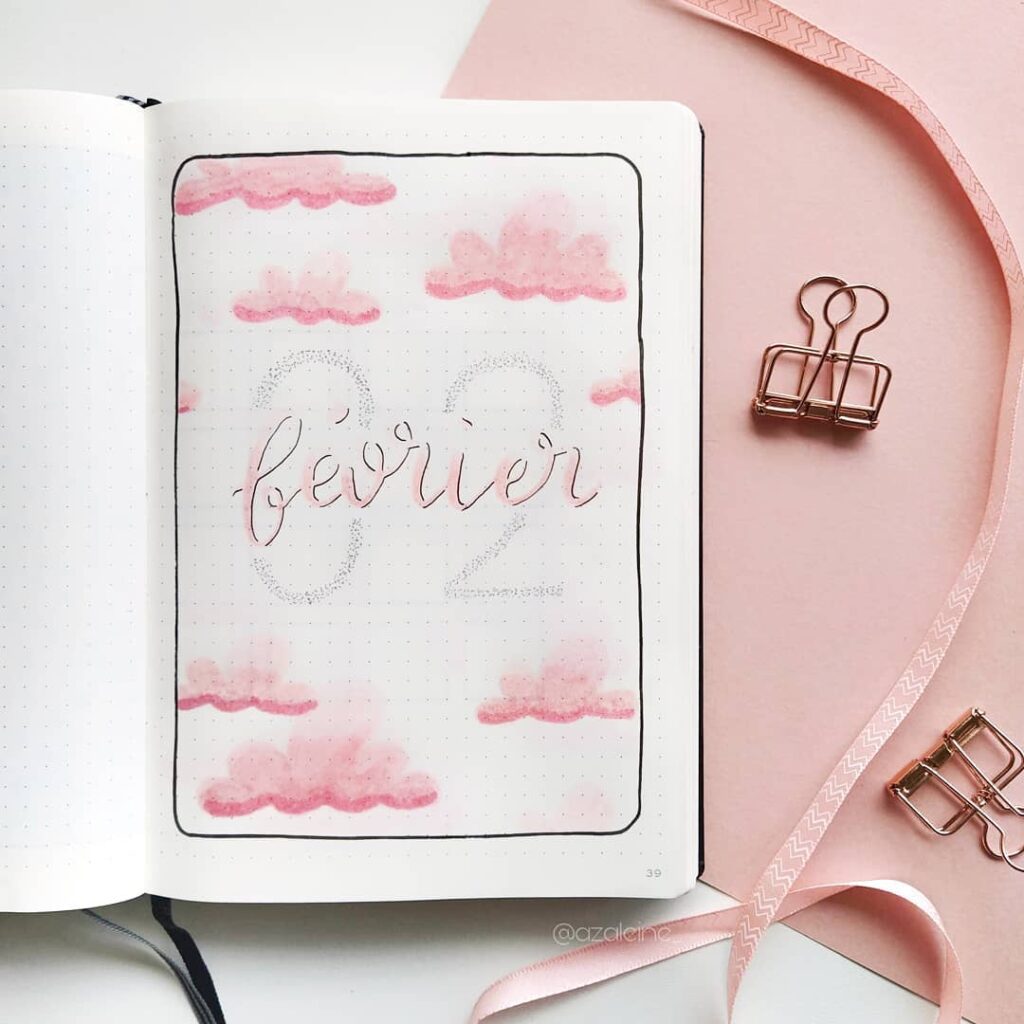 Bullet journal February cover with pink clouds and decorative script text on dotted paper, surrounded by pink stationery.
