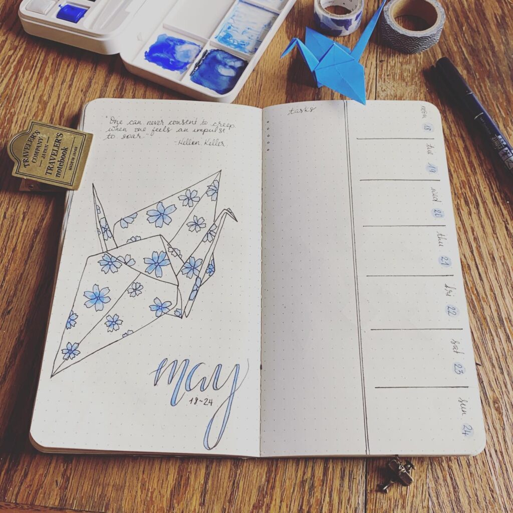 Open bullet journal with origami crane and watercolor palette on a wooden table.