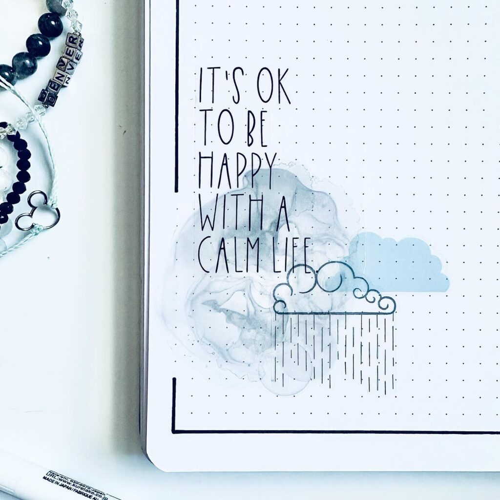 Notebook page with the text It's OK to be happy with a calm life, accompanied by cloud and rain illustrations.