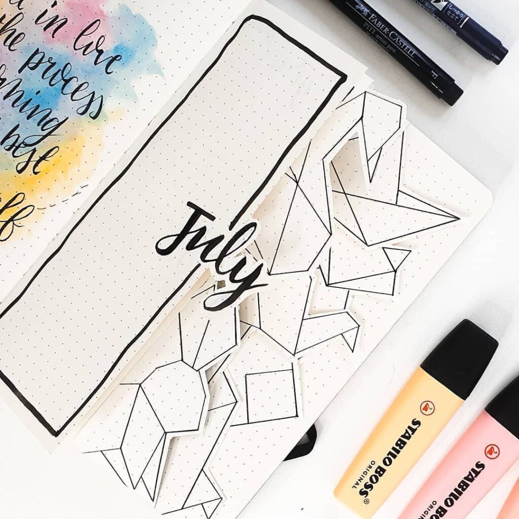 Bullet journal open to July page with geometric designs, colorful pens, and motivational quote in calligraphy.
