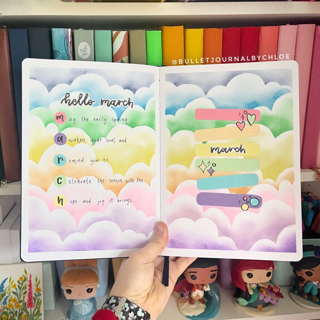 Colorful bullet journal spread for March with pastel clouds and creative headers, surrounded by books and figurines.