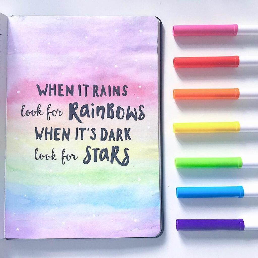 Inspirational rainbow quote on pastel notebook page beside colorful markers on white background.