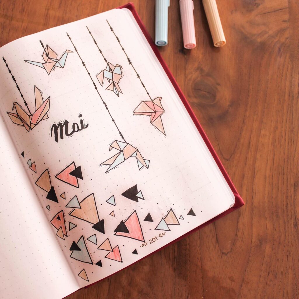Bullet journal with origami bird doodles and Mai text, surrounded by pastel pens on a wooden table.