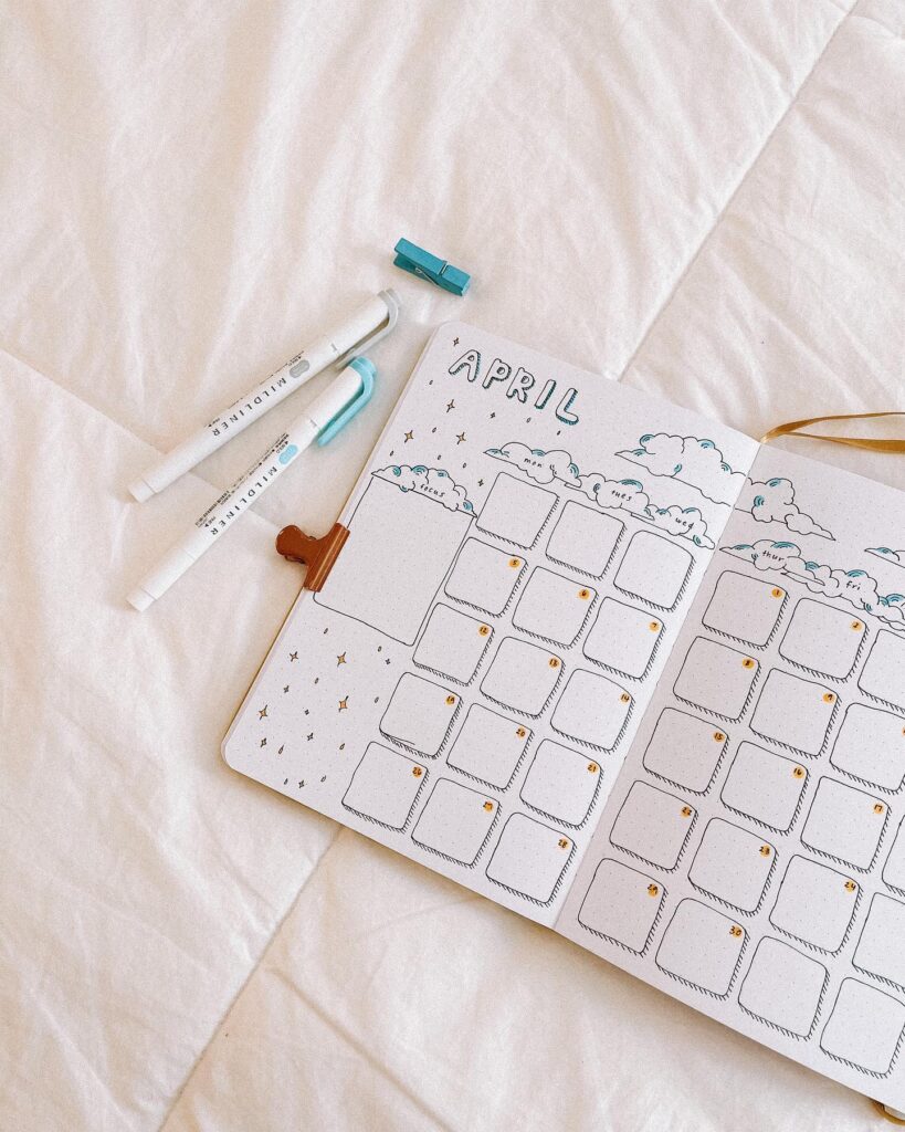 April bullet journal layout with clouds, displayed on a white bed, alongside pens and a clip for creative planning.