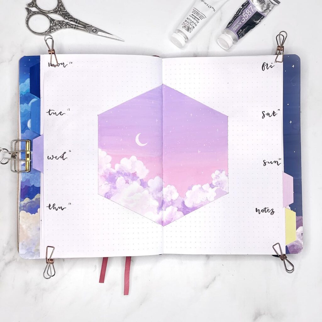 Open bullet journal with a pastel purple sky and clouds illustration, surrounded by art supplies.