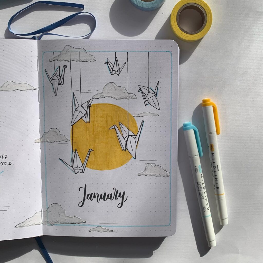 Bullet journal January cover with origami cranes, clouds, and a sun, surrounded by washi tape and pens.