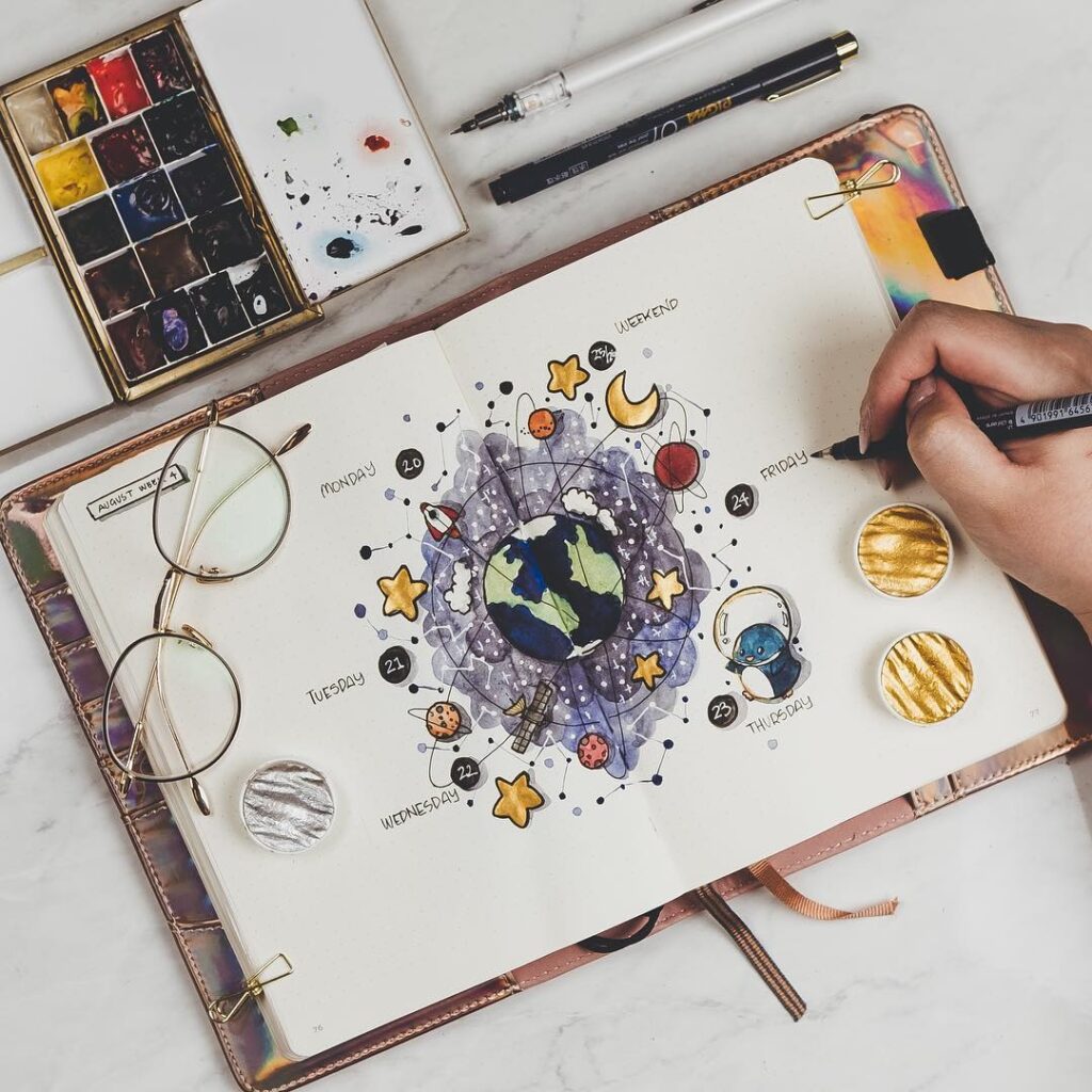 Hand drawing celestial-themed journal entry with watercolor paints and glasses on a desk.