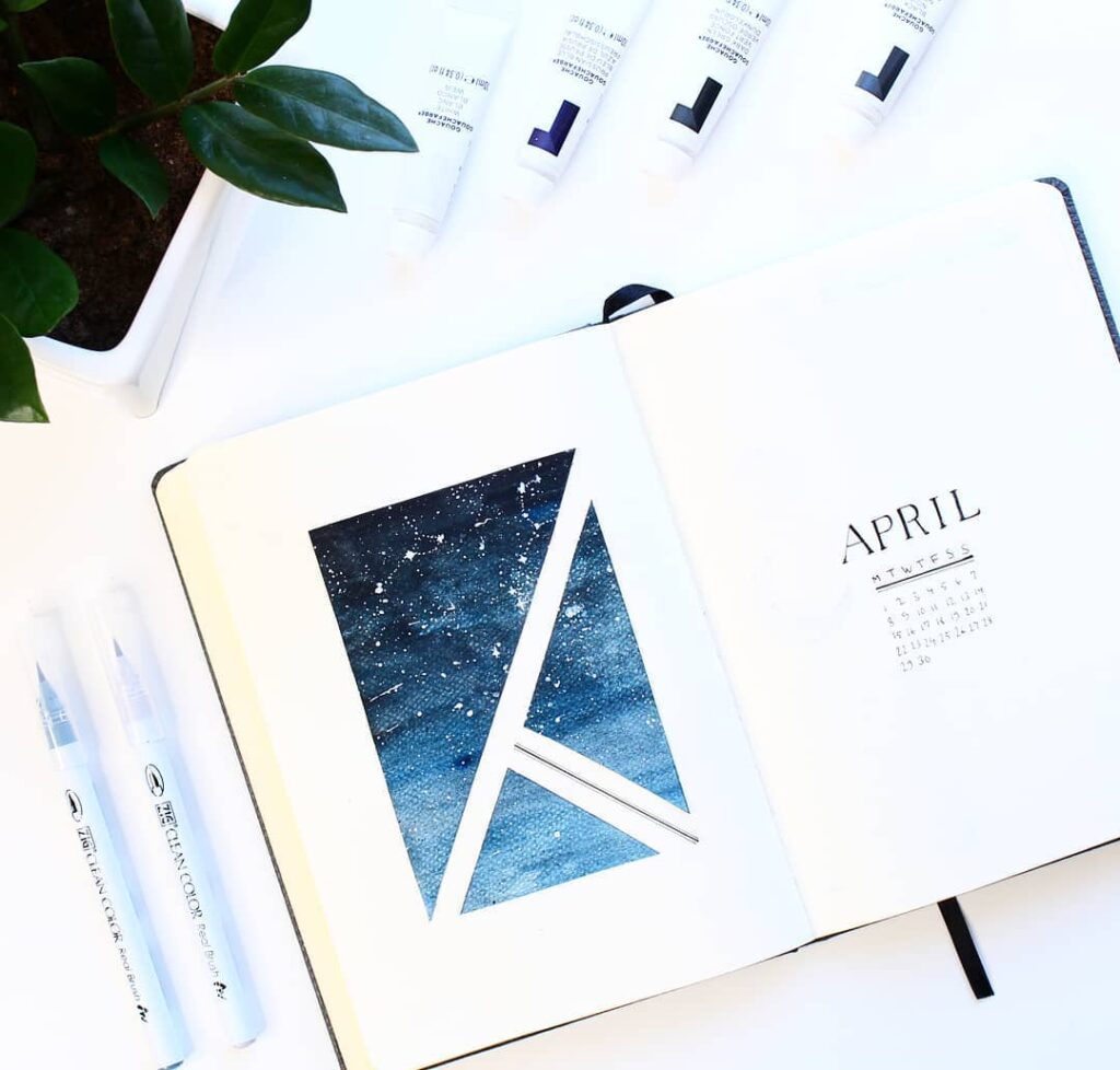 Open notebook with geometric galaxy art and April calendar, surrounded by pens and plant on white background.