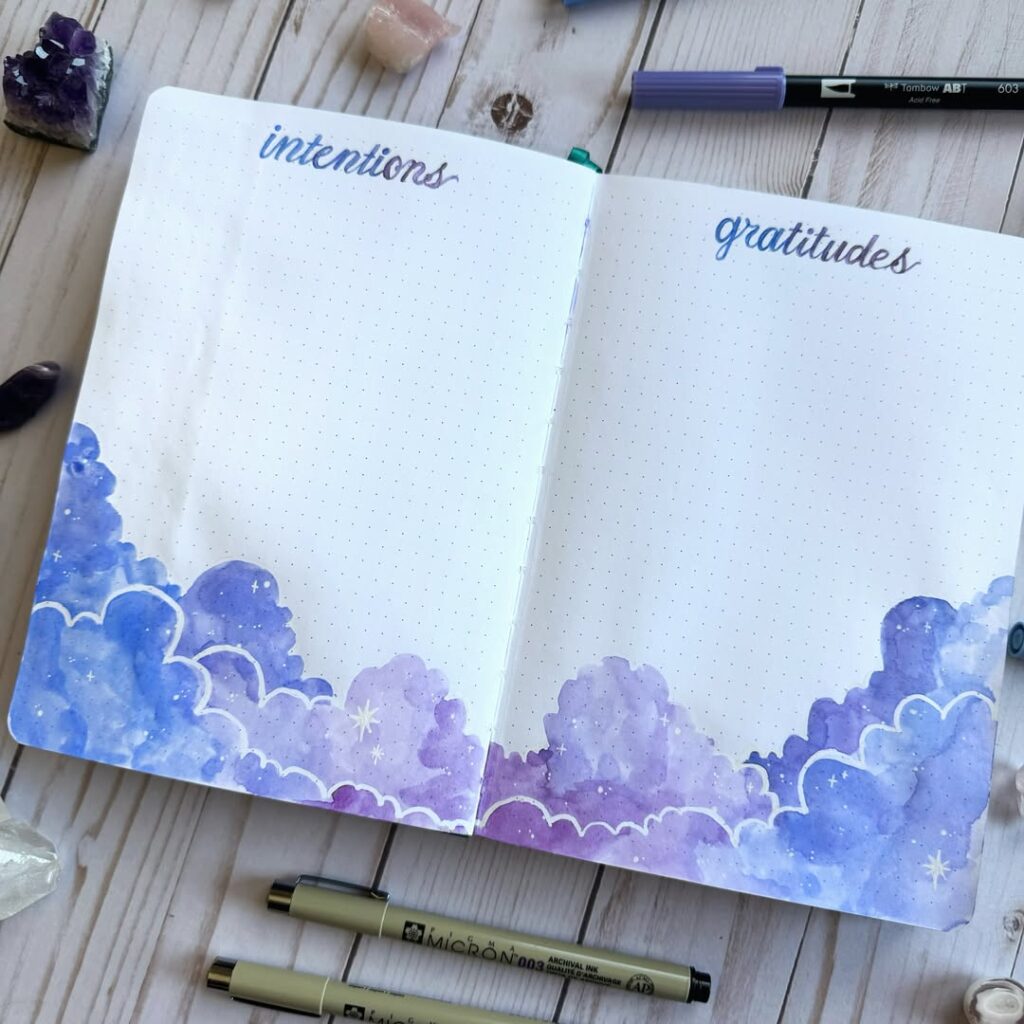 Open journal with intentions and gratitudes pages, surrounded by purple watercolor clouds and pens on a wooden table.