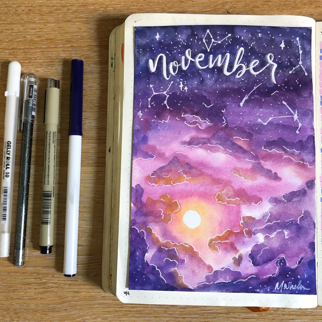 Watercolor journal page for November with purple sky, stars, and sunrise design, alongside pens on a wooden table.