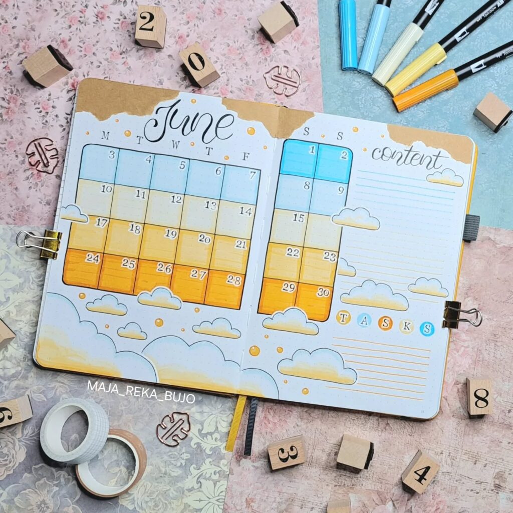 June bullet journal spread with colorful calendar, clouds, and task section, surrounded by markers and stamps.