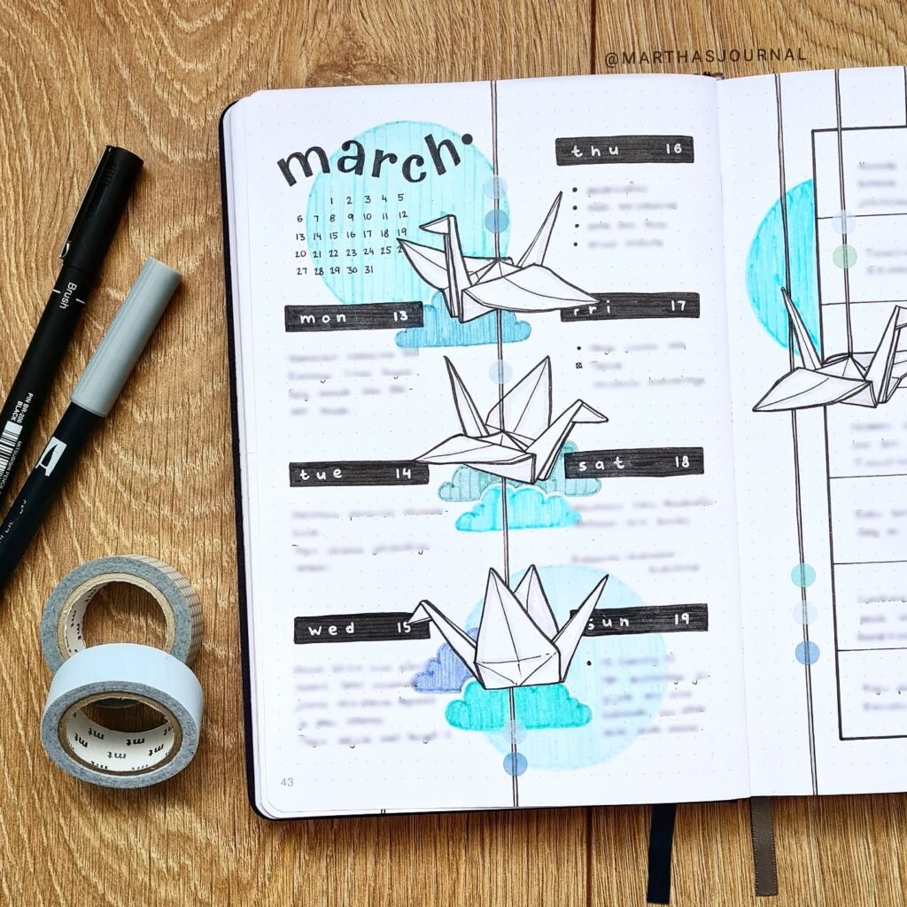 March bullet journal spread with origami cranes and blue clouds, featuring pens and washi tape on a wooden surface.