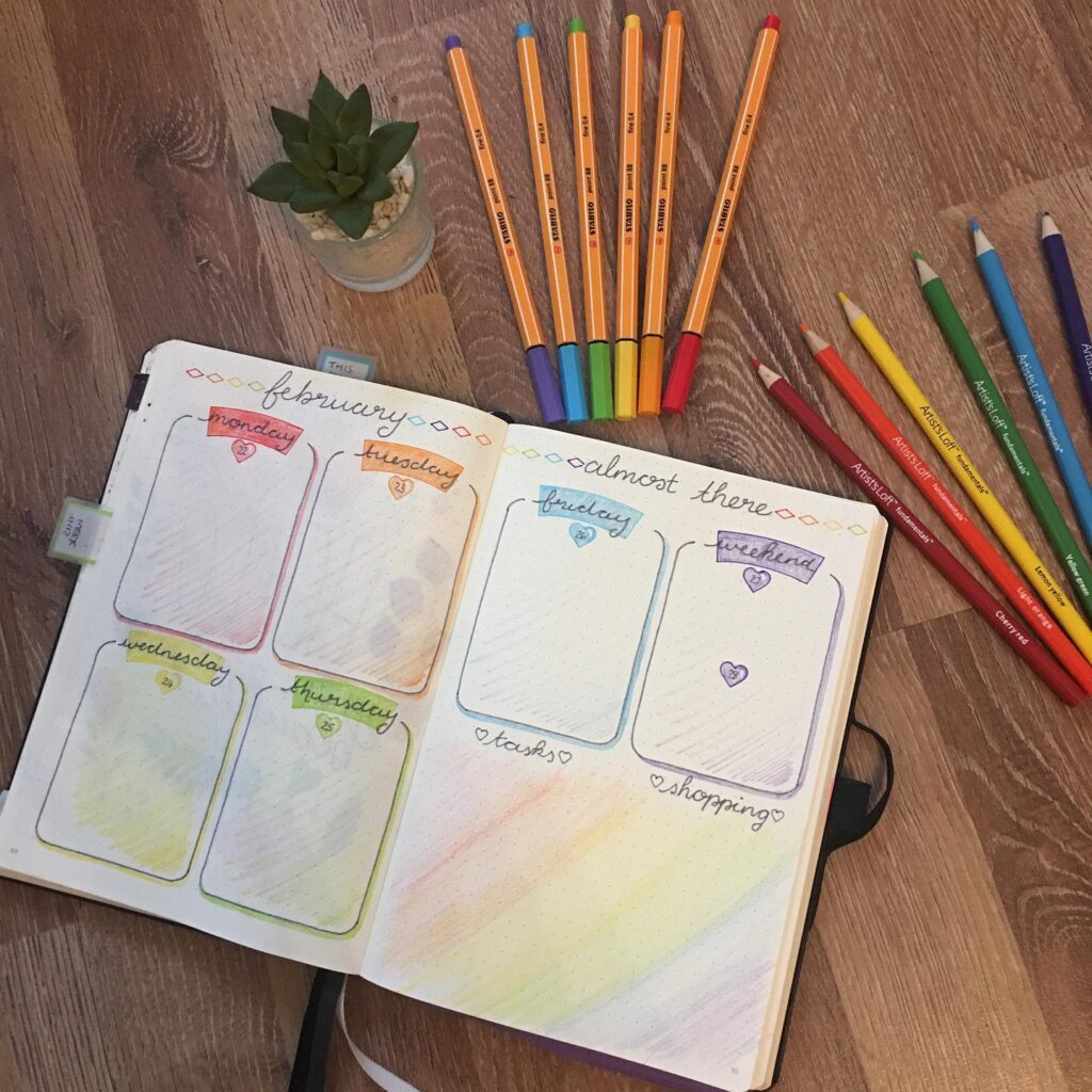 Colorful bullet journal with weekly planner, surrounded by pens, pencils, and a small plant on a wooden floor.
