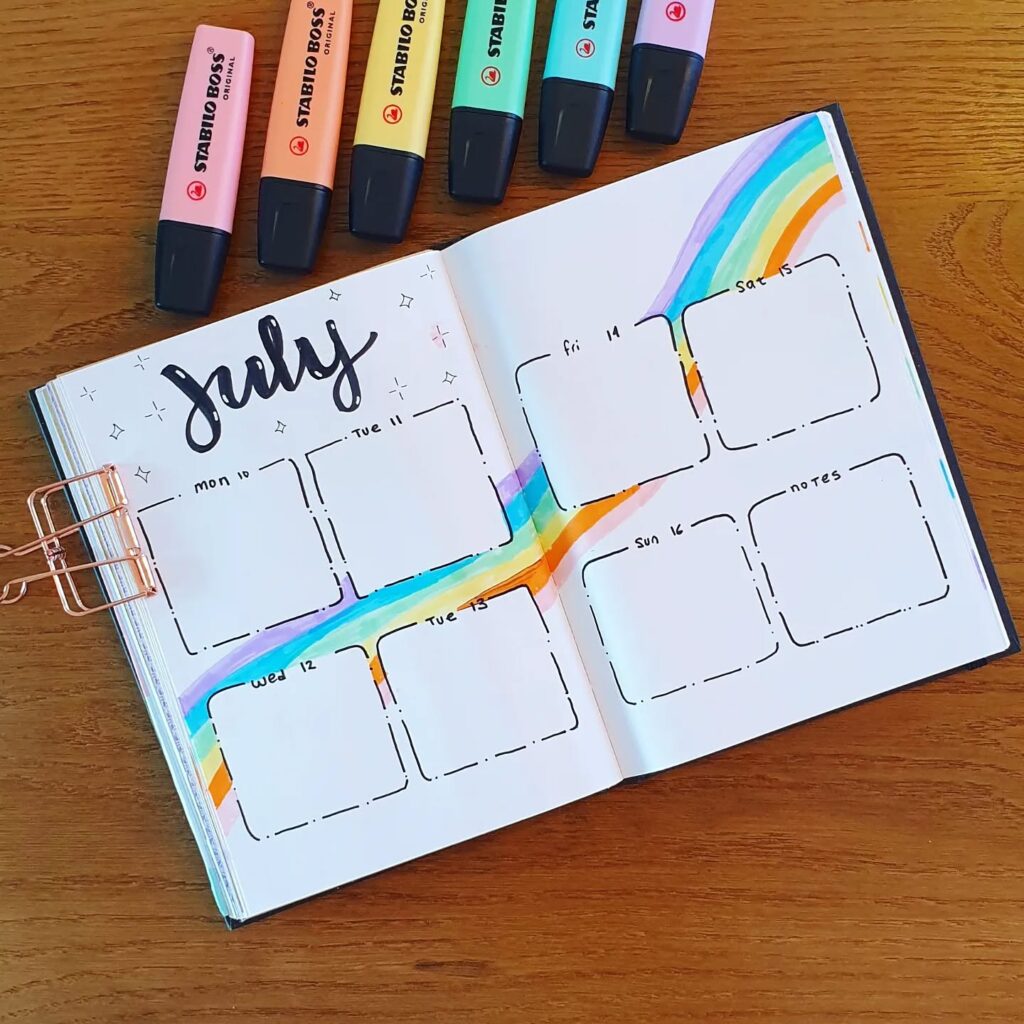 Open bullet journal for July with rainbow design, surrounded by colorful Stabilo Boss highlighters on wood table.