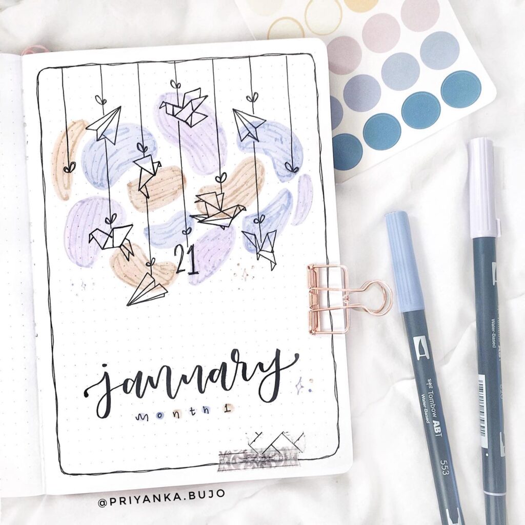 Bullet journal January cover with hand-drawn paper airplanes and pastel colors, featuring pens and a sticker sheet.