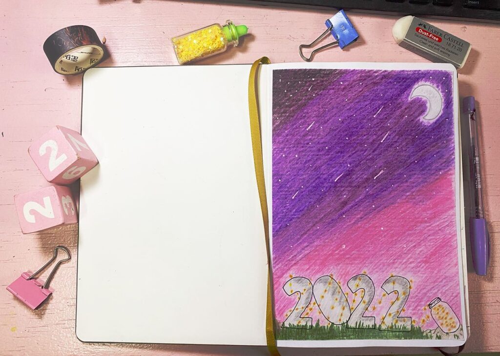 Open notebook with colorful 2022 drawing on pink desk. Blocks, glitter, and stationery surround.