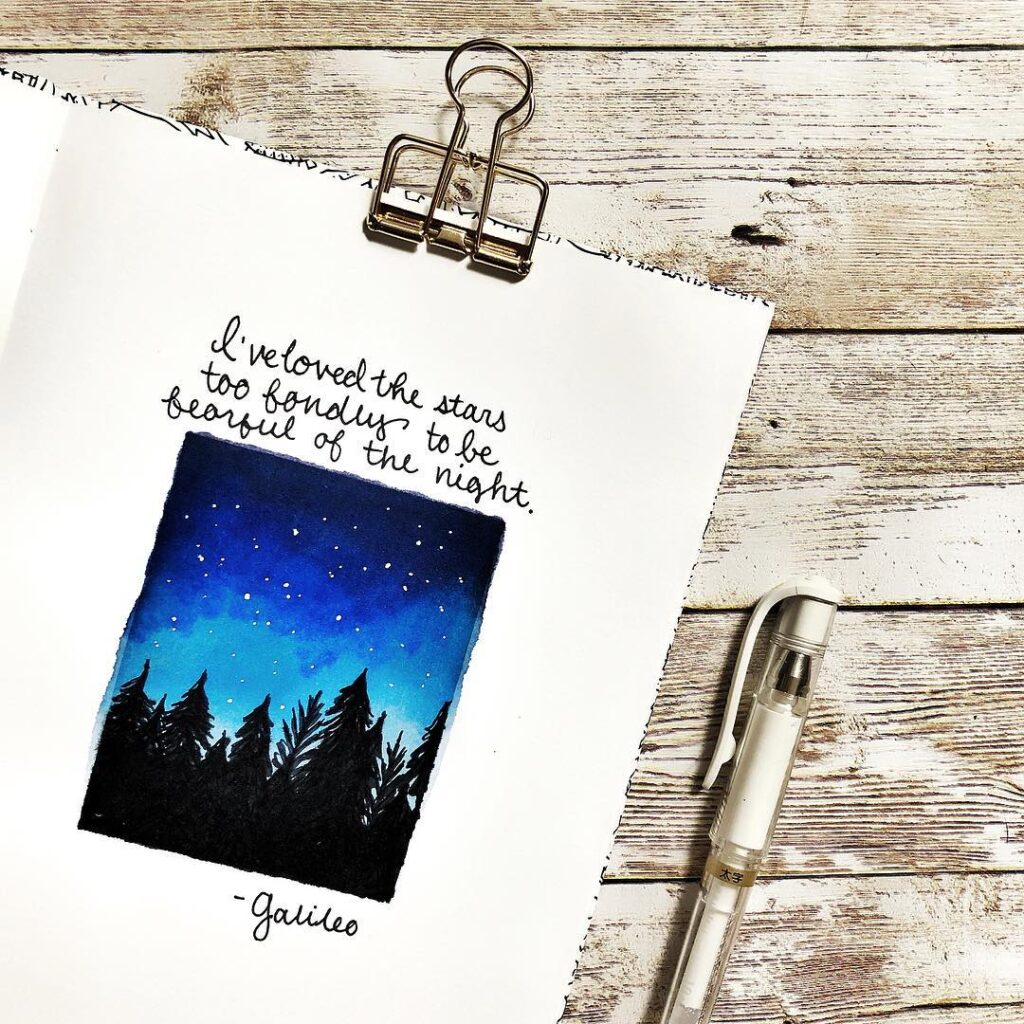 Inspirational quote on a notebook page with a night sky illustration next to a white pen on wooden background.