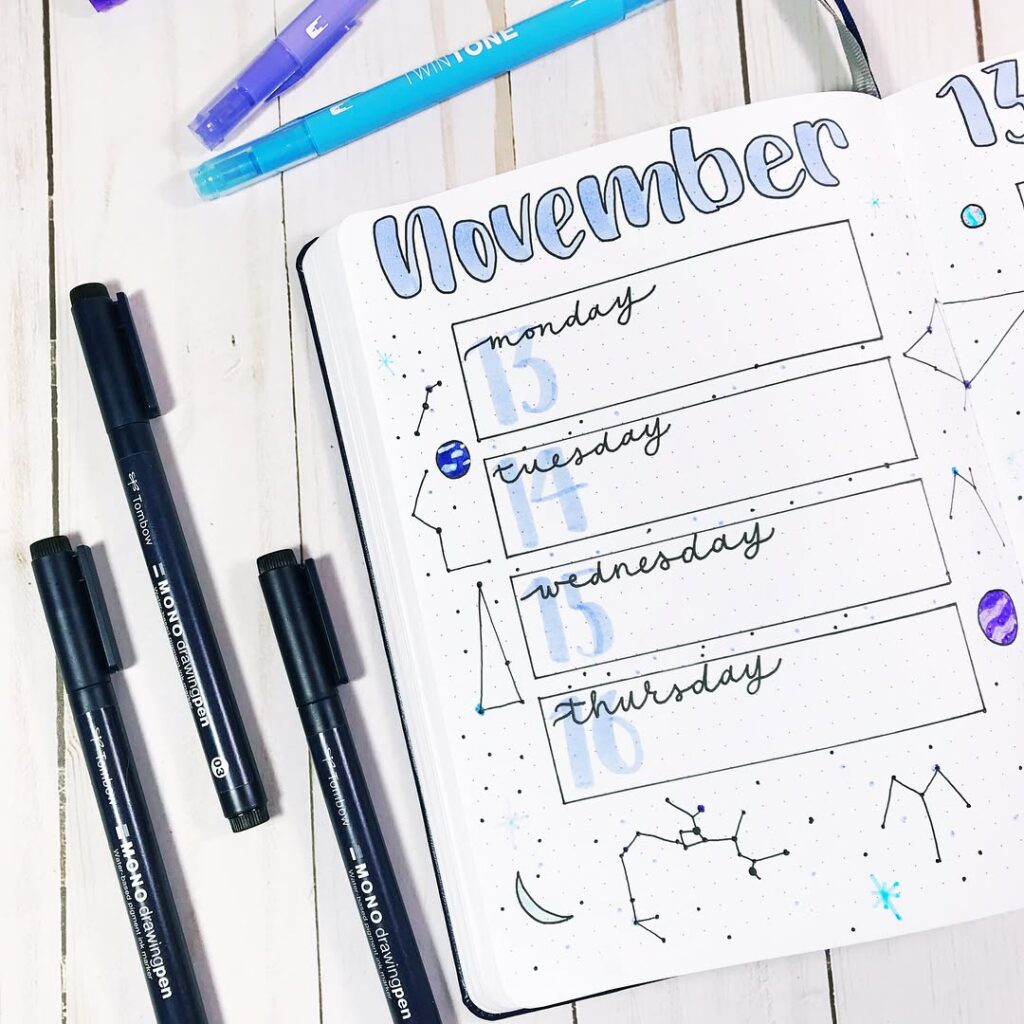 Bullet journal with November weekly layout, featuring blue and black pens on a white desk.