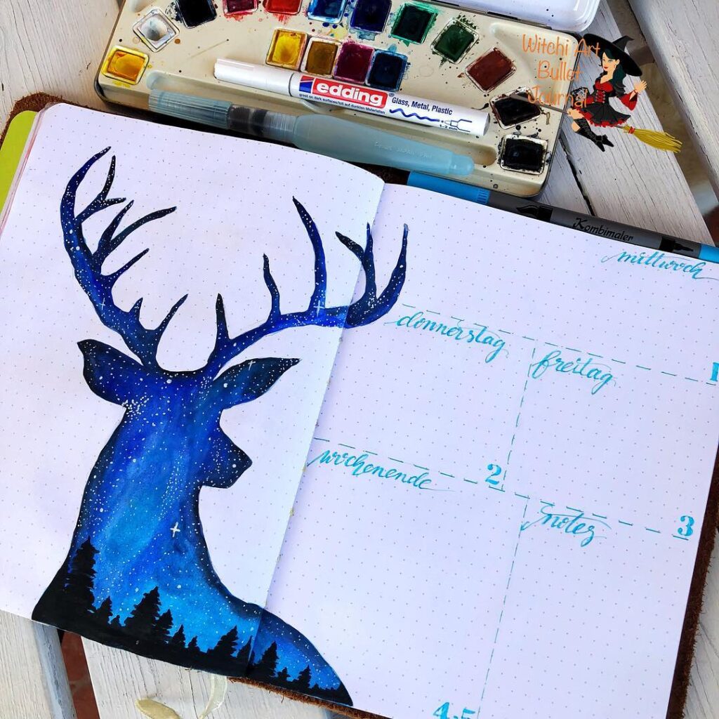 Bullet journal with watercolor deer art and weekly planner layout next to paints and pens.