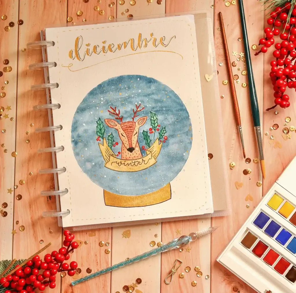 Festive December journal with a winter reindeer design, surrounded by paintbrushes and red berries on a wooden table.